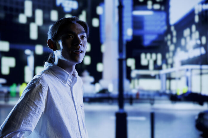 Asian man wandering around city boulevards during nighttime, being amazed by nightlife environment buildings. Citizen strolling around empty avenues at night, enjoying metropolitan skyscrapers